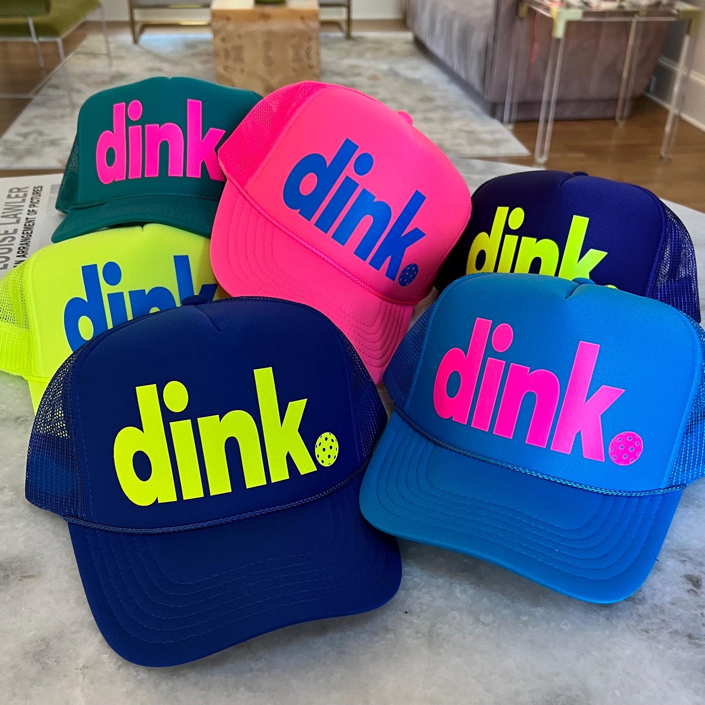 Five trucker hats with mesh back panels. The hats are in various colors including teal, pink, yellow, blue, and dark blue. The hats all have the word 'dink' written on the front in a bold, neon colored font. The word 'dink' is in a different color on each hat. The hats are arranged in a semi-circle on a light-colored surface. The period is a pickleball perfect for playing!