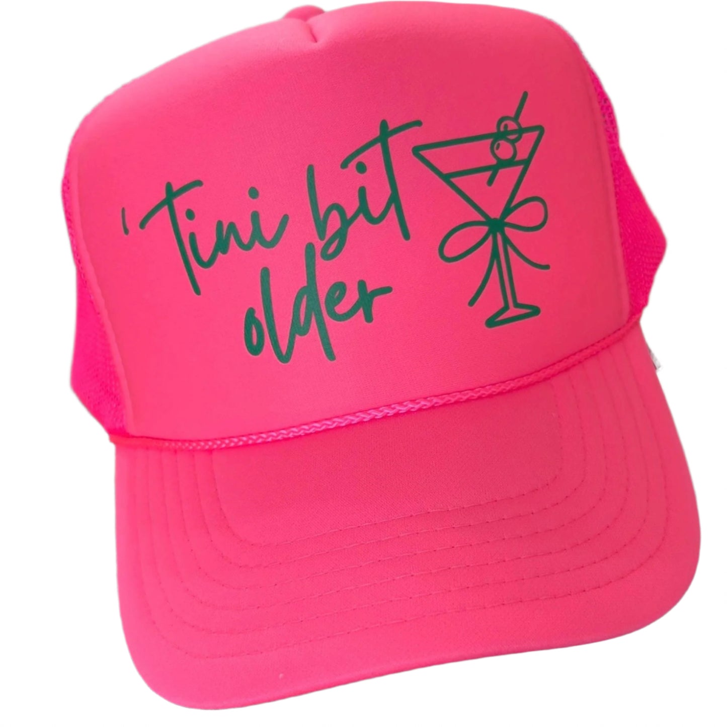 Neon pink trucker hat with green ink that says "tini bit older"