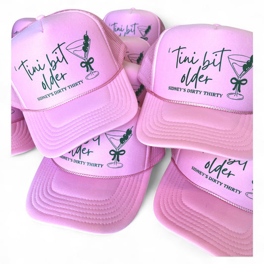 Stack of light pink foam trucker hats with martini glass graphic and “‘Tini Bit Older – Sidney’s Dirty Thirty” text, custom 30th birthday party caps.

