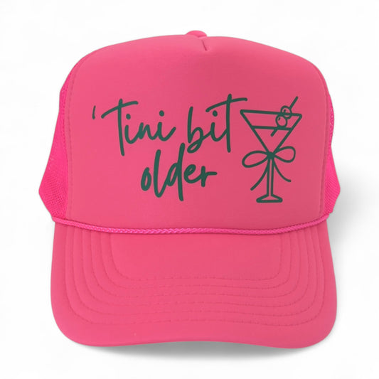 Neon pink foam trucker hat with green script “‘Tini bit older” and martini glass graphic, fun birthday party hat.
