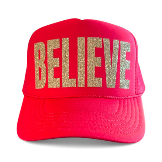 Santa inspired red Christmas trucker hat with 'BELIEVE' in gold glitter on a white background. 