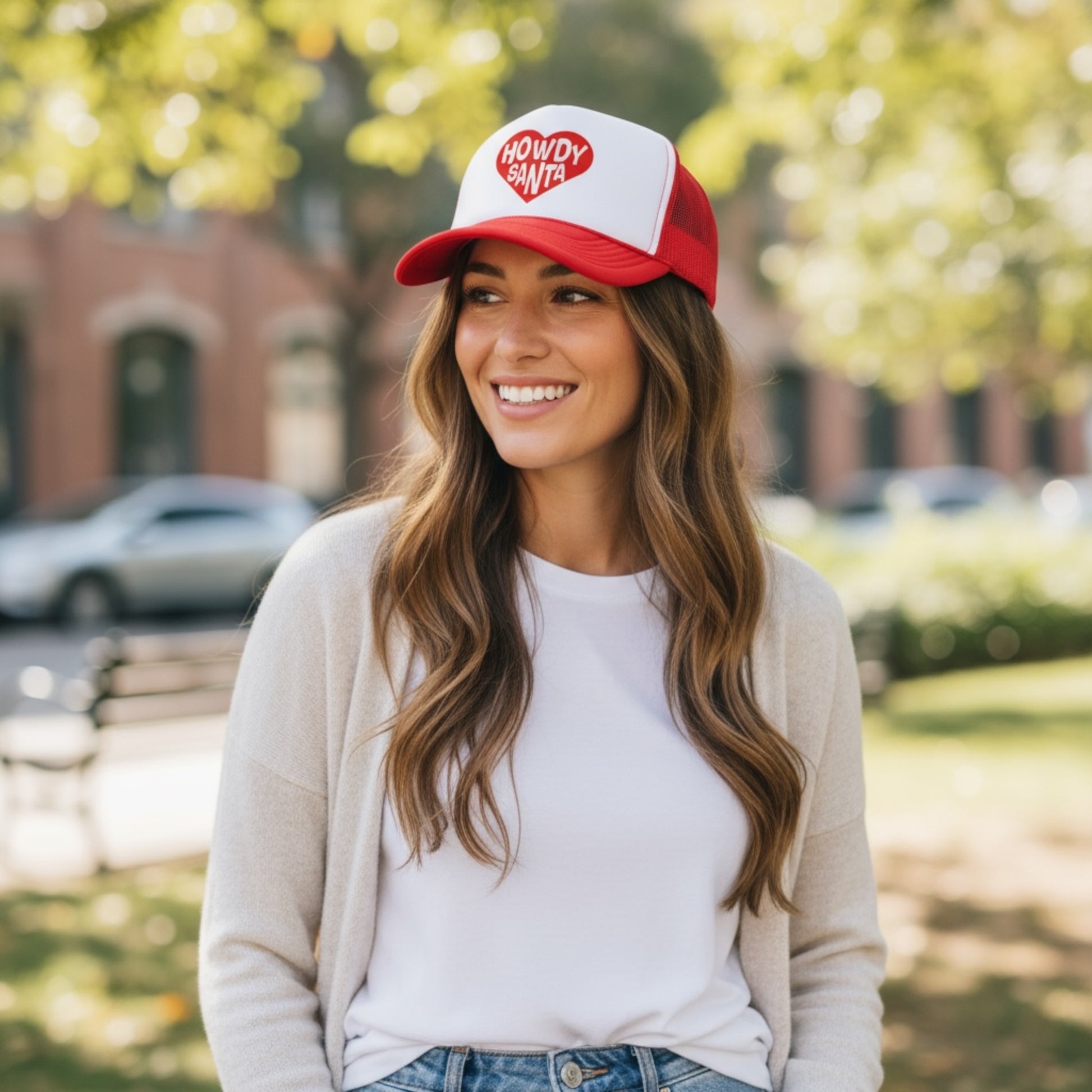 Woman in casual outfit wearing red and white “Howdy Santa” Christmas trucker hat outdoors in a sunny park.
