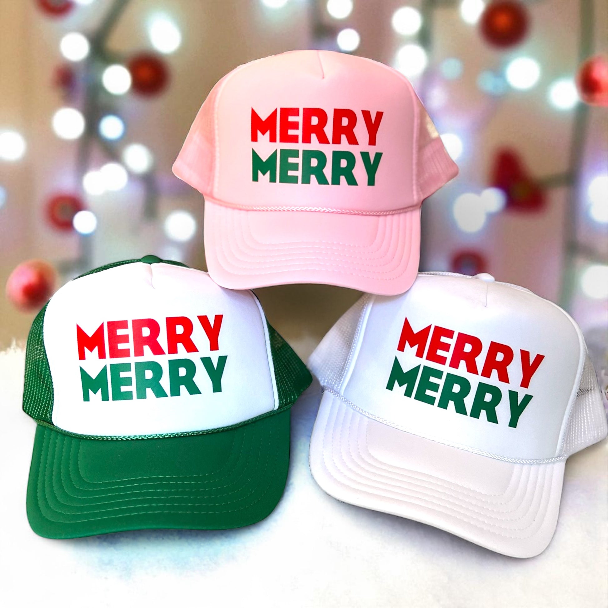Three “MERRY MERRY” festive Christmas trucker hats in pink, green, and white displayed in front of blurred holiday lights and ornaments. Perfect gift for the holidays!